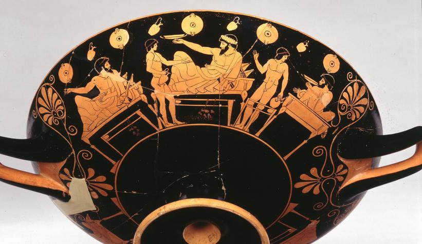 Tre græske mænd ligger på kliner ved et græsk symposion. De holder en drikkeskål hver, en kylix. Og to unge drenge er klar til at hælde mere i drikkeskålene. På væggen hænger drikkekopper og kander. C. 480 f.Kr.